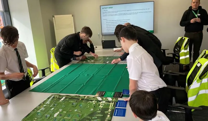 A group of school-age students in uniform lean over a large table, working on an interactive activity.